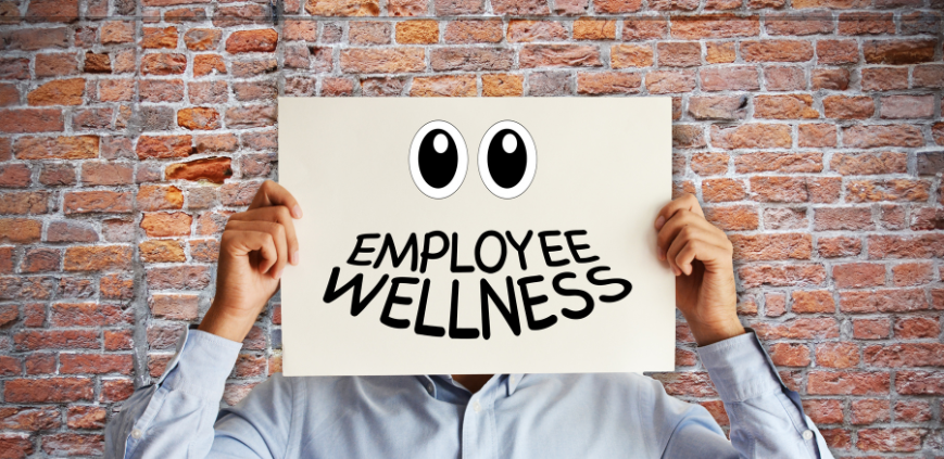 A man holding a large sign over his head that says Employee Wellness against a brick wall.