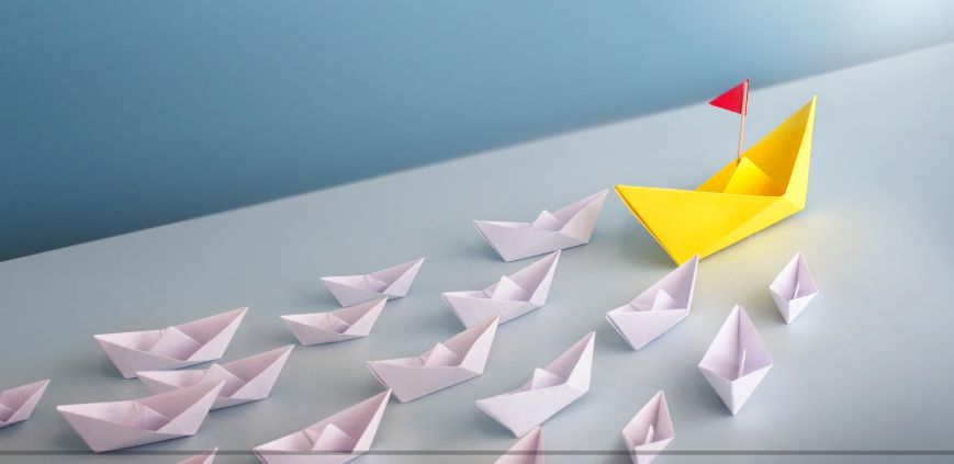 White paper boats in multiple lines behind a large yellow paper boat with a red flag, set against a light blue background.