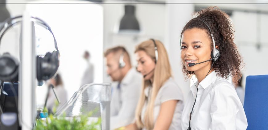 A woman wearing a headset sitting in front of a computer in a call center, with other employees in the background.