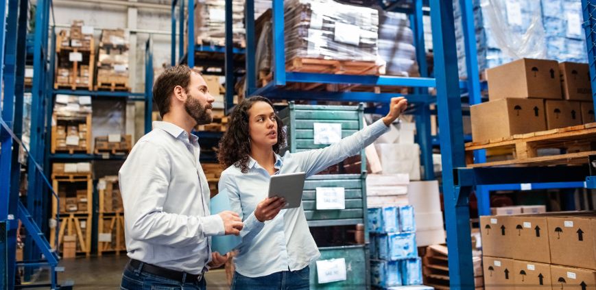 A man and woman in a warehouse looking at inventory. The woman is holding a clipboard.