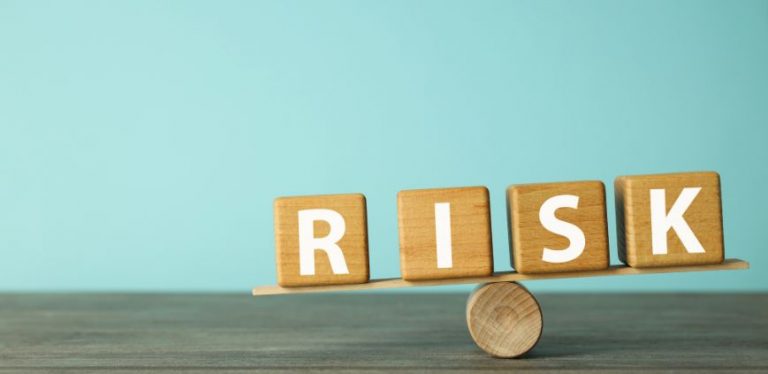 Wooden blocks that spell RISK balancing on a wooden dowel against a blue background.